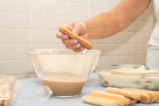 A close-up of a hand dipping a ladyfinger into coffee, preparing a classic tiramisu dessert in a kitchen