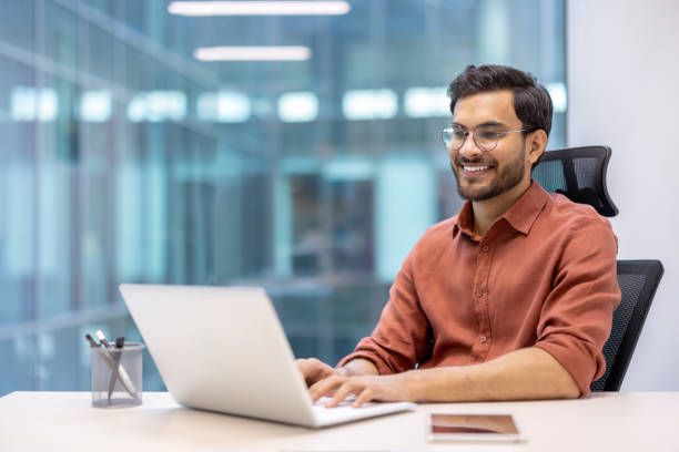 istock Smiling professional man using laptop at modern office workstation with smartphone on desk 2167823087