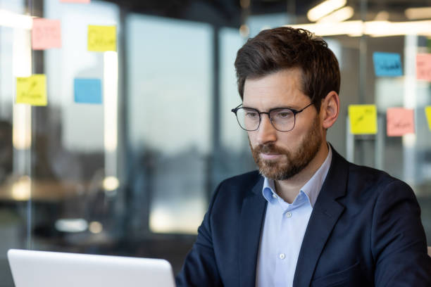 istock Focused businessman working on laptop in modern office with notes on glass wall 2167824641