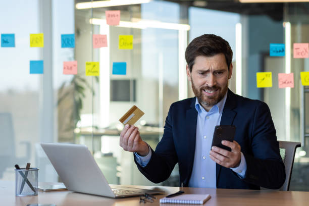 istock Concerned businessman holding credit card while looking at smartphone in modern office environment with sticky notes on glass wall 2167825251