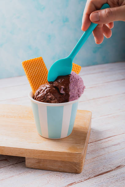 istock Hand holding plastic colored spoon on ice cream scoops topped with chocolate sauce and waffer cookies. 2167829794