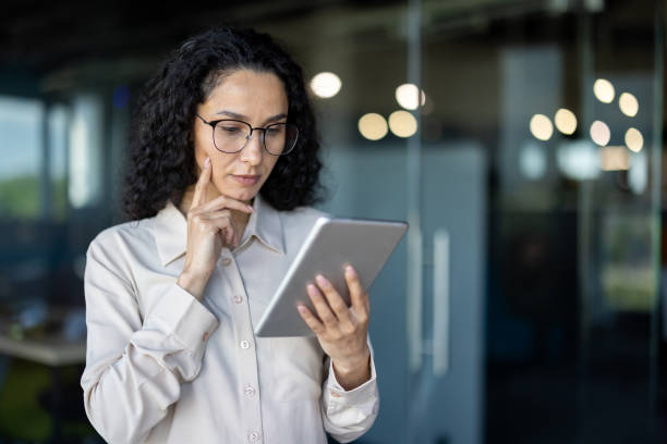 istock Serious confident thinking business woman using tablet computer, financier looking at company reports online, woman boss in shirt and glasses standing near window, Hispanic woman with curly hair 2167830113