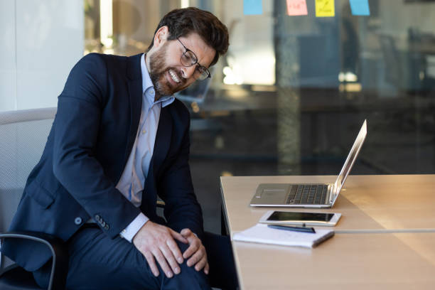 istock Businessman experiencing leg pain while sitting at desk in modern office space 2167834132