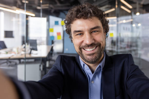 istock Friendly businessman in office during video call, smiling and looking at camera, using phone for a professional meeting, showcasing modern workspace in corporate setting. 2167834554
