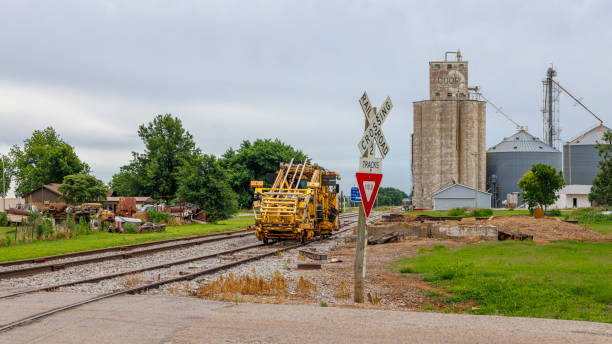 Greenfield, Blaine County, Oklahoma, USA - May 30, 2024: Yellow railroad maintenance vehicle positioned on tracks at rural railway crossing. Large grain silos and storage tanks dominate background, indicating agricultural area. Railroad crossing sign visible in foreground