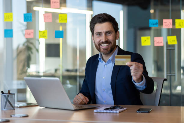 istock Smiling businessman holding a credit card while working on laptop in office 2167837409