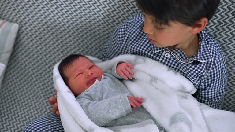 Older brother lovingly holding newborn sibling, both gazing at each other. Heartwarming moment of sibling bonding and family connection during infancy and childhood