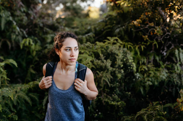 An active and adventurous woman of Eurasian descent looks to her side while walking through lush plants during a hike on a beautiful day.