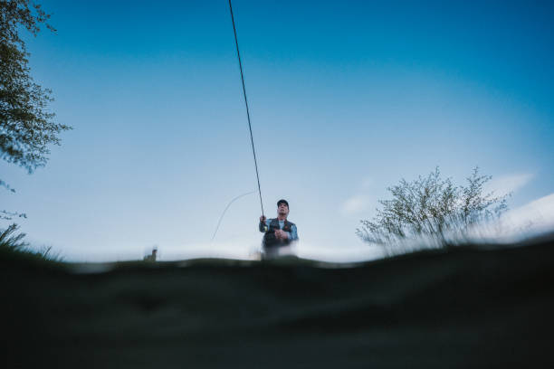 Low angle view looking up at a Caucasian male fisherman stanings on the bank of the Deschutes River while fly fishing, shot partially underwater.