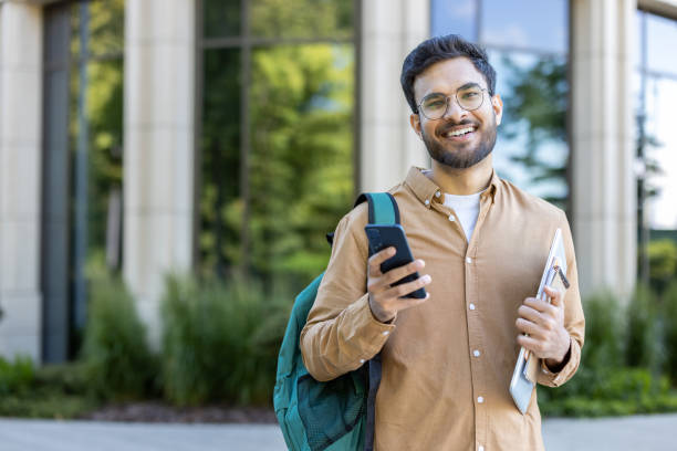 istock Happy Indian student with phone and backpack standing outside campus building 2167841628