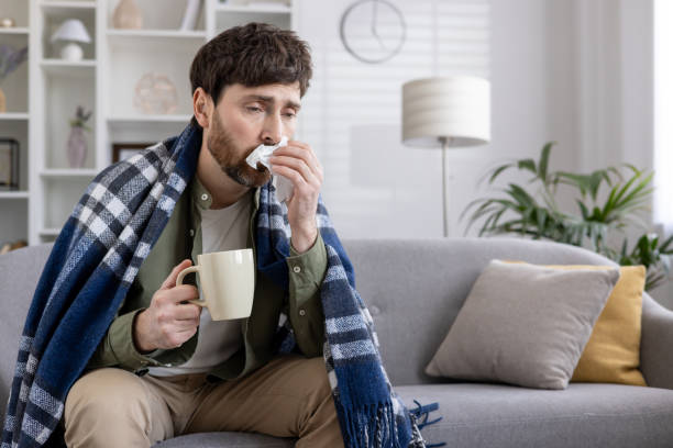 istock Sick man resting on couch with blanket and hot drink at home, blowing nose with tissue 2167853343