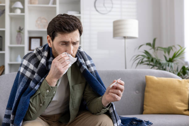 istock Man sick with flu sits on sofa holding tissue and thermometer covered with blanket 2167855918