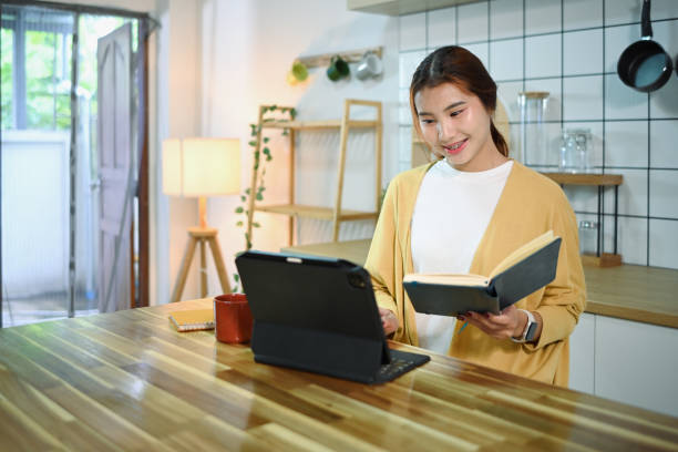 istock Smiling young asian woman holding book using digital tablet on kitchen counter 2167858857