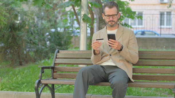 istock Young Man Shopping Online with Phone while Sitting Outdoor 2167859030