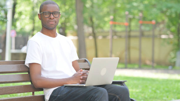 istock Young African Man Looking at Camera while Using Laptop Outdoor 2167859656