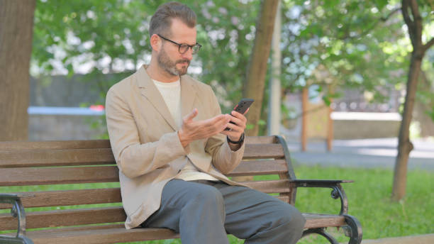 istock Middle Aged Man Upset by Loss on Phone while Sitting Outdoor on Bench 2167860356