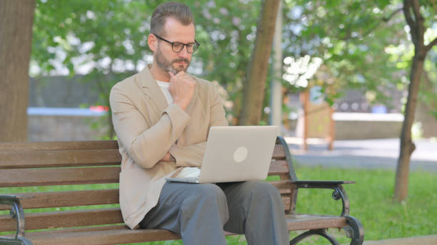 istock Middle Aged Man Brainstorming Middle Aged Man Reading on Laptop while Sitting Outdoor on Bench 2167861351