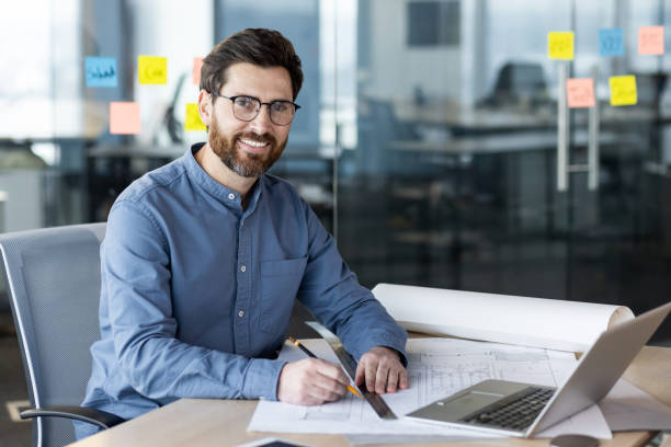 istock Smiling architect working on blueprints at office desk with laptop and sticky notes 2167862237
