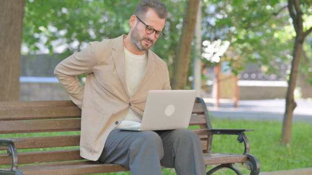 istock Middle Aged Man with Back Pain Using Laptop while Sitting Outdoor on Bench 2167862737