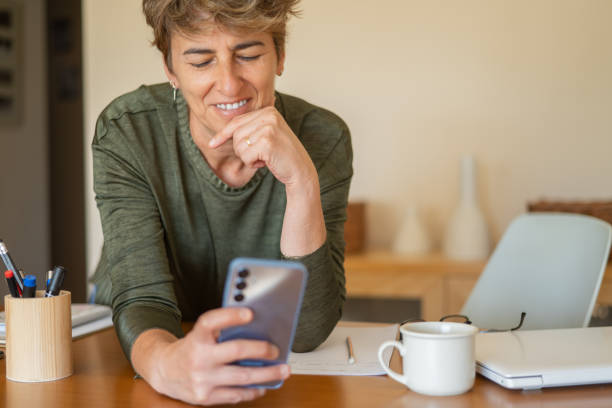 Smiling Mature Woman Using Smartphone At Workplace Or Home, Good News, Enjoying Exchanging Messages, Distracted From Work, Spending Time On Internet, Chatting With Friends Or Family, Watching News. mujer madura sonriente usando un teléfono inteligente en el lugar de trabajo o en casa, buenas noticias, disfrutando del intercambio de mensajes, distraída del trabajo, pasando tiempo en internet, chateando con amigos o familiares, viendo noticias.