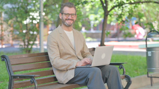 istock Middle Aged Man with Laptop Smiling at Camera while Sitting Outdoor on Bench 2167864720