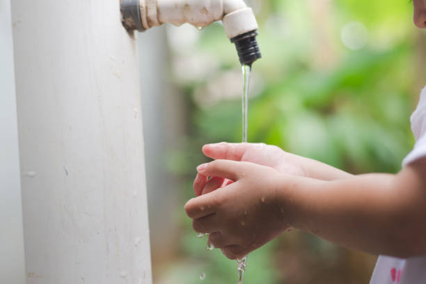 A young girl stands at an outdoor tap, fascinated by the gentle stream of water flowing over her hands. Captures the wonder and joy of a child discovering the simple joys of nature in a peaceful garden