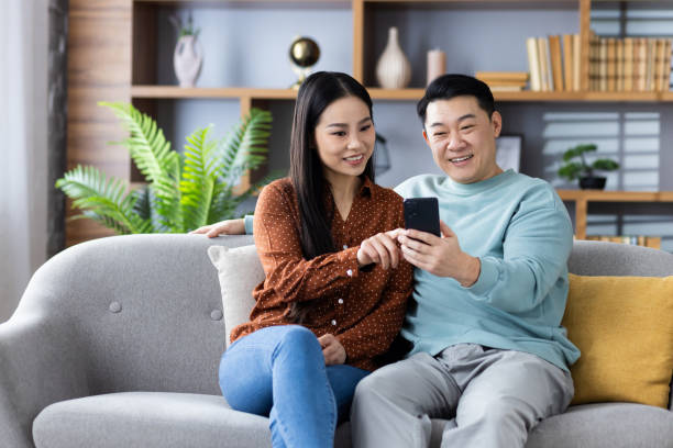 istock Asian couple sitting on couch using smartphone, browsing internet together 2167870745