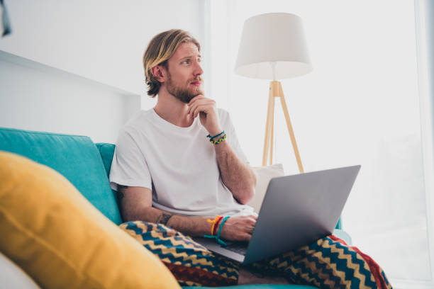 istock Photo of handsome young guy sit couch netbook skeptical think dressed nightwear spend pastime spacious house indoors room 2167886544