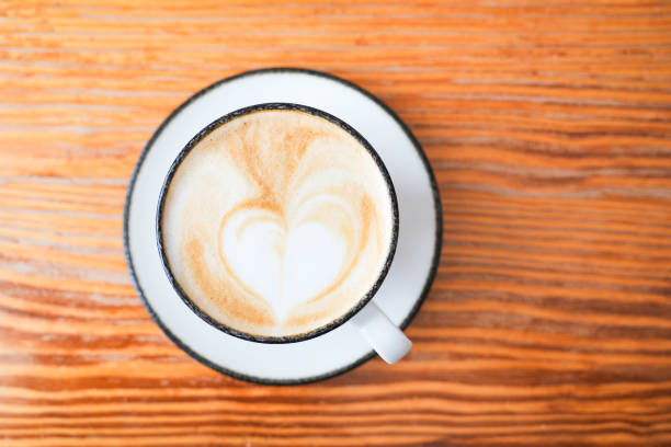 A white coffee cup with a heart design on it sits on a wooden table. The cup is filled with coffee, and the heart design adds a touch of warmth and affection to the scene