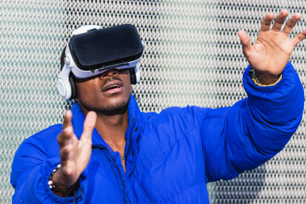 African American male standing on street near metal fence while exploring virtual reality in innovative goggles and touching air