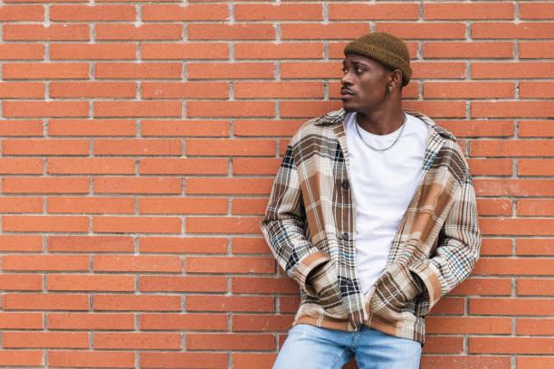 Young African American guy in checkered shirt and trendy hat frowning and looking away while standing against brick wall on city street