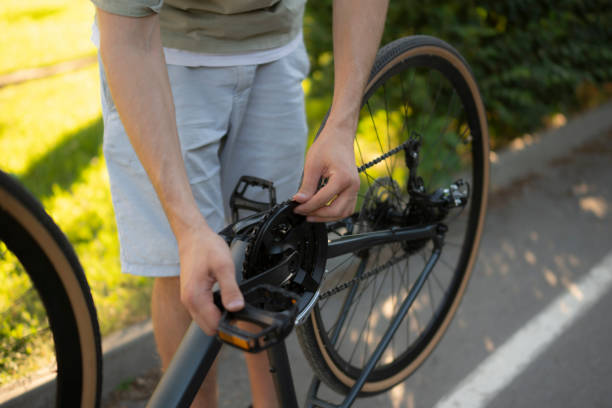 istock man adjusts the wheel of a bicycle while standing on a paved path. Green grass and trees are in the background 2167967986