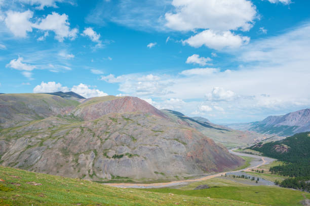 Mountain river flows around sunlit big multicolor hill under clouds in blue sky. Alpine river surrounds large mountain of many vivid colors in changeable weather. Shadows of clouds in high mountains.