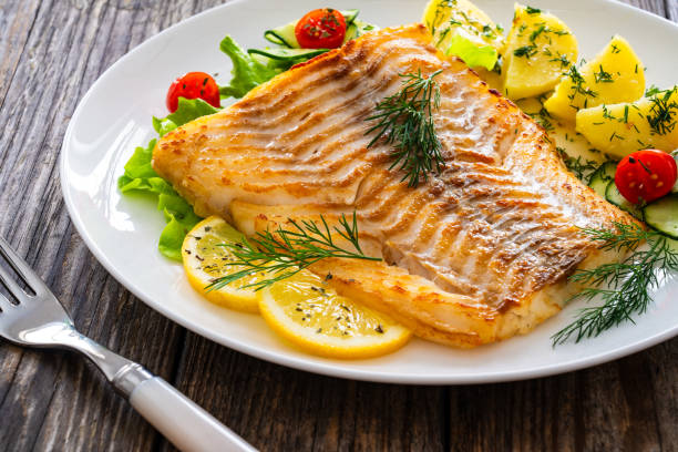 istock Fried cod fillet, boiled potatoes, fresh vegetables and lemon slices served on white plate on wooden table 2167995606