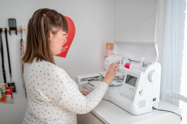 A woman operates an embroidery machine in her well-lit home studio, adjusting the settings as she begins her crafting project. She is wearing a star-patterned sweater, and the scene is filled with natural light, creating a focused and serene environment for her creative work.