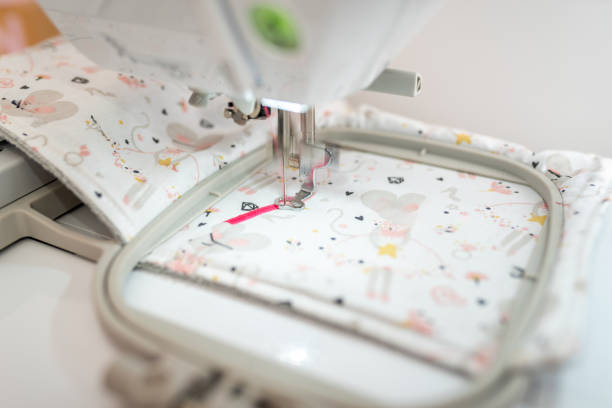 A close-up view of an embroidery machine in action, stitching with pink thread on a piece of patterned fabric. The fabric features whimsical designs, and the precision of the embroidery process is highlighted, capturing the detailed work involved in crafting.