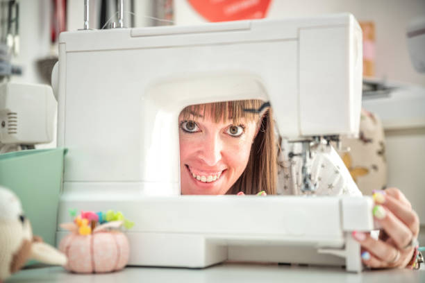 A woman is playfully peeking through the arm of a sewing machine, smiling brightly. The sewing machine, along with other crafting tools and decorations in the background, creates a fun and engaging atmosphere in a well-organized and creative workspace.