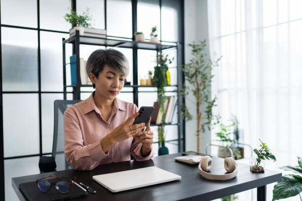 istock A female architect in a light pink shirt uses her smartphone while sitting at her desk, which is neatly arranged with various work tools, in a bright and modern office environment 2168012388