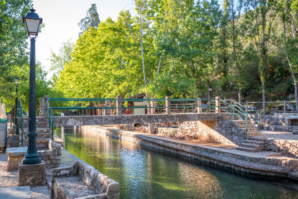 A picturesque view of a stone bridge spanning a peaceful canal in the village of Alte, Algarve. The scene features lush green trees, a traditional streetlamp, and rustic stonework, reflecting the charm and natural beauty of this historic Portuguese village under a clear, sunny sky.