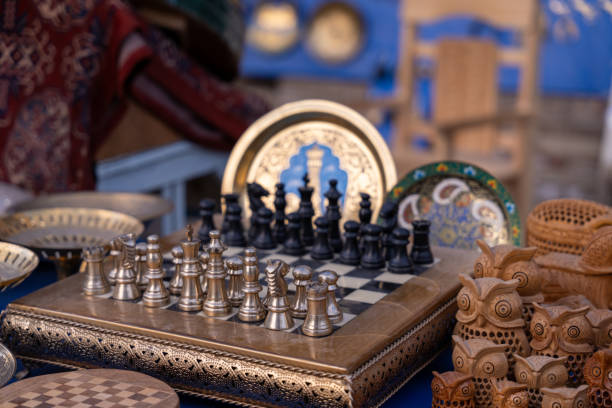 istock A chess set is displayed on a table with other items. The chess set is made of metal and has a wooden base. The table is covered with various items, including a chair, a bowl, and a vase 2168019866