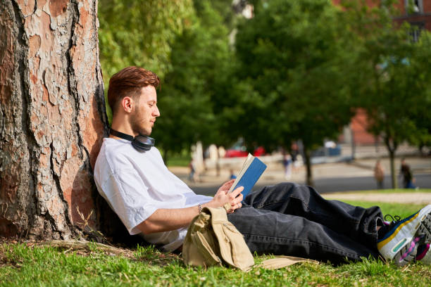 istock A man is sitting under a tree reading a book. He is wearing headphones and has a backpack next to him. The scene is peaceful and relaxing, with the man enjoying his time outdoors 2168019900