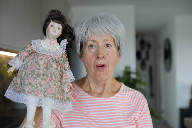 A gray haired Caucasian senior woman is holding an interesting porcelain doll. The doll has pale complexion and could be really creepy.