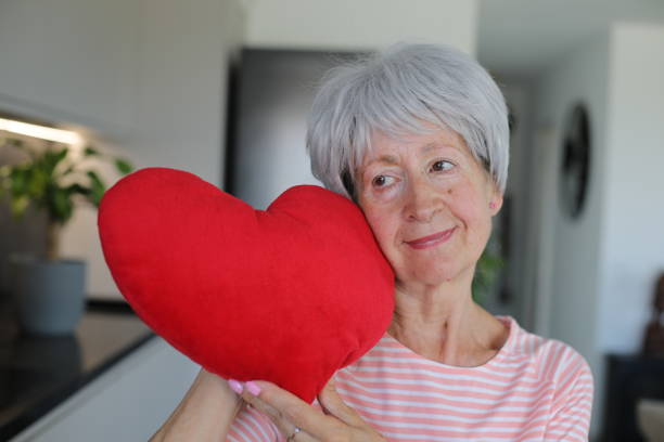 A really pretty senior Caucasian woman in her late sixties is holding a large stuffed heart. She has short gray hair and looks very natural.