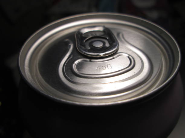 Close-up of a hand holding an opened soda can on a gravel path. A colorful wristband and sneakers are visible, capturing a casual outdoor moment.