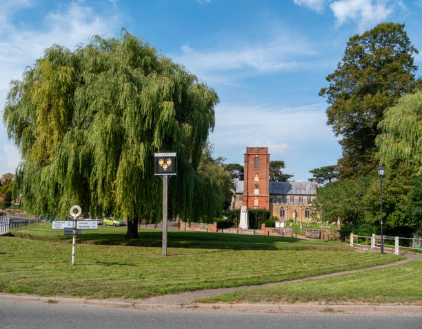 Seen across the village green in Grundisburgh, Suffolk, Eastern England, is St Mary’s church, its churchyard, village sign and war memorial. The church shows evidence from various centuries, starting in the 14th, while its red brick tower dates from the early 18th century: it is known for its beautiful angel hammerbeam roof. The River Lark flows across the green.
