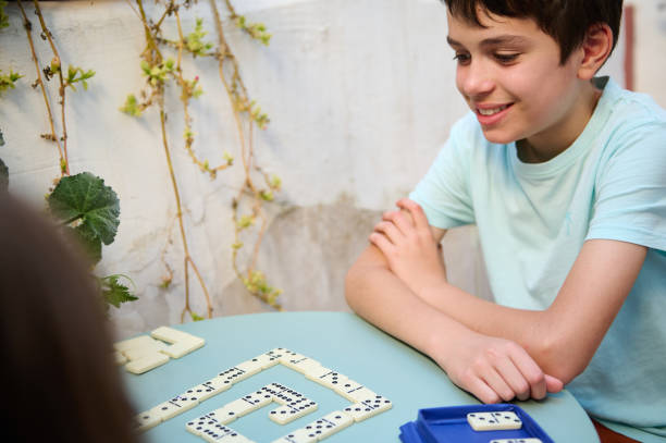 istock Smiling boy playing dominoes at a table outdoors, enjoying a fun and engaging game with friends or family in a sunny garden setting, showcasing leisure and happiness. 2168059062