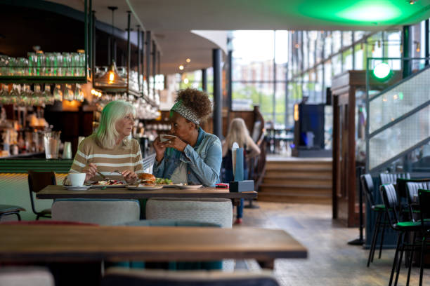 A full shot of two mature female friends catching up with each other sat in a bar located in Newcastle Upon Tyne. They are both having brunch together laughing and talking with each other. \n\nVideos are available similar to this scenario