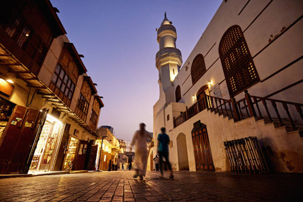 Cobblestone walkway through historic Jeddah at dusk Surface level view of Saudi male pedestrians, illuminated storefronts, and minaret. jeddah stock pictures, royalty-free photos & images