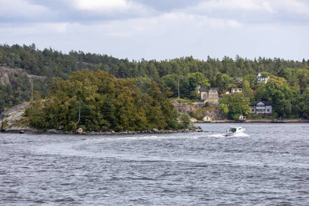 A scenic view of a small island with lush greenery and a boat cruising by in the foreground. The background features a shoreline with charming houses and trees under a cloudy sky.