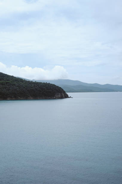 Elevated coastal view on Kınalıada showing a rocky foreground, clear blue water with surface ripples, a headland forming a sheltered bay, and a working boat offshore beneath a bright sky.